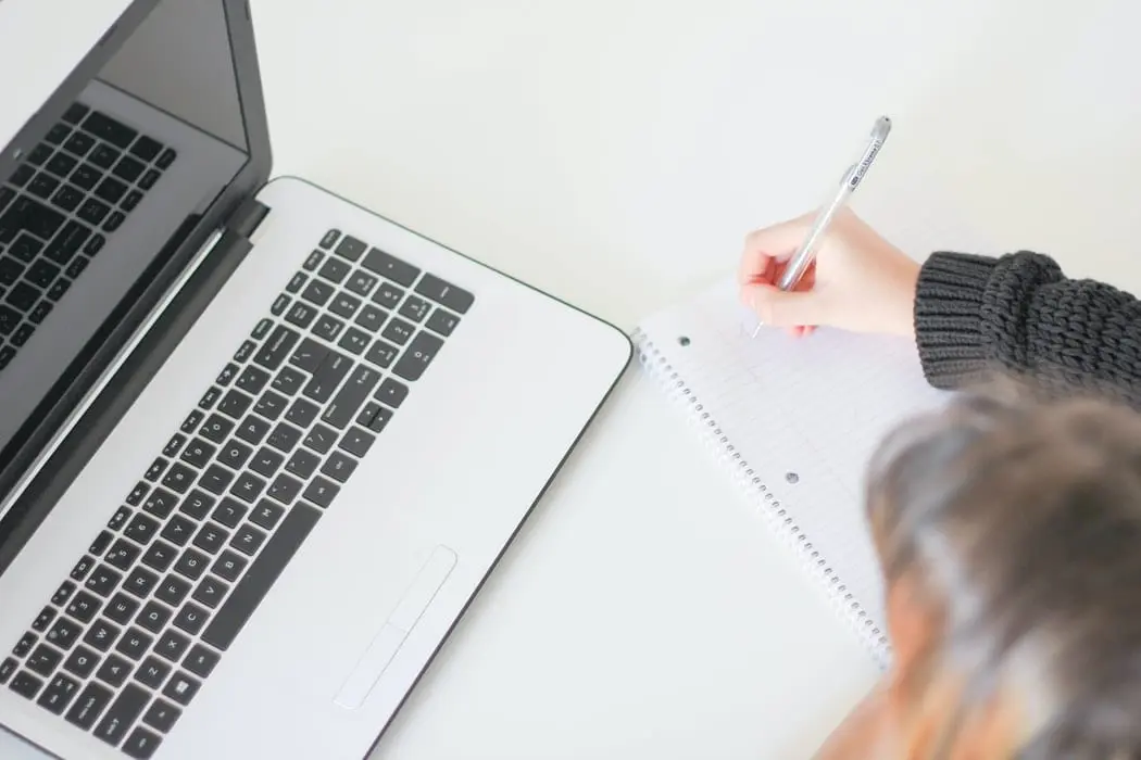 Person writing in a notebook with a laptop sitting on the table.