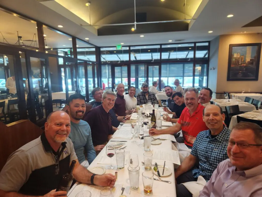 A group of thirteen men smile and pose for a photo around a long, set dining table in a well-lit restaurant, holding menus and drinks, with large windows in the background.
