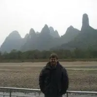 A person wearing a dark jacket stands outdoors in front of a railing, with misty, jagged mountains and greenery visible in the background under an overcast sky.