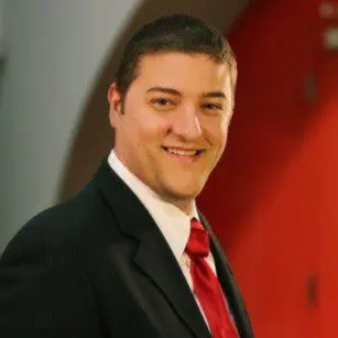 A man in a dark suit, white shirt, and red tie smiles at the camera. He has short dark hair and is standing in front of a blurred, red and gray background.