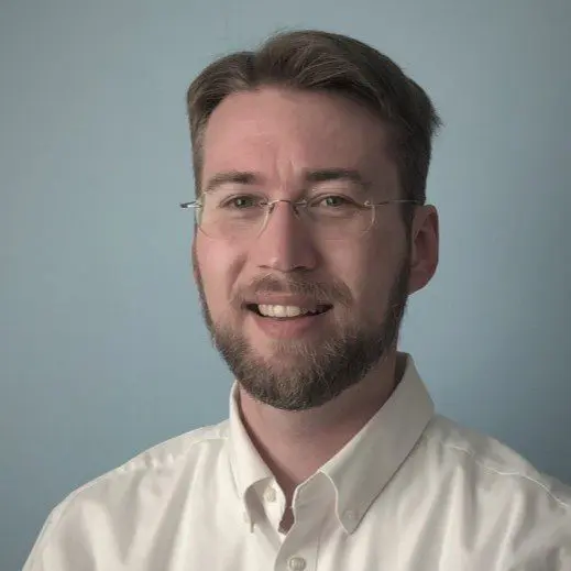 A man with short brown hair, a beard, and glasses is smiling at the camera. He is wearing a white collared shirt and standing against a plain light blue background.