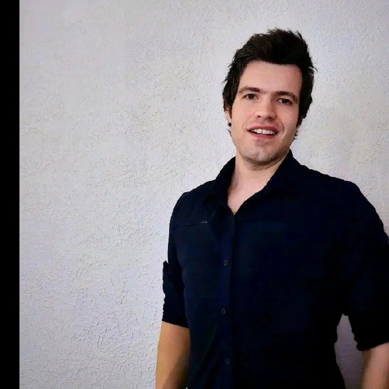 A young man with short, dark hair, wearing a dark button-up shirt, stands smiling in front of a plain, light-colored textured wall.