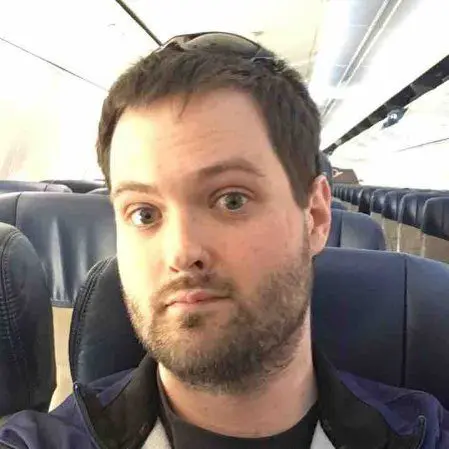 A man with short brown hair and a beard sits in an empty airplane cabin, looking at the camera with a neutral expression. He wears a navy jacket and sunglasses resting on his head.