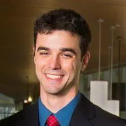 A man with short dark hair, wearing a dark suit, blue shirt, and red tie, smiles at the camera. The background is indoors with modern lighting and a wooden ceiling.