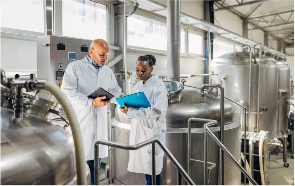 Two scientists in white lab coats review notes on tablets and clipboards inside an industrial laboratory with large stainless steel tanks and technical equipment in the background.