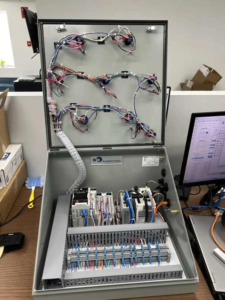 An open electrical control panel box shows organized wiring, circuit boards, relays, and terminals inside. The panel is on a desk near a computer monitor and office supplies. The box lid is propped open, displaying internal components.