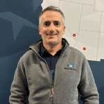 A man in a gray zip-up jacket stands smiling in front of a blue wall with a partial map of the southwestern United States and red location markers.