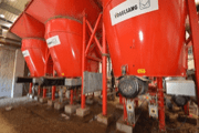 Large red feed silos with metal legs and motors are installed inside a barn, raised above the ground on concrete blocks. The silos are used for storing and distributing animal feed.