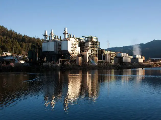 An industrial facility with large white structures and smokestacks is reflected in a calm body of water, surrounded by hills and trees under a clear blue sky.