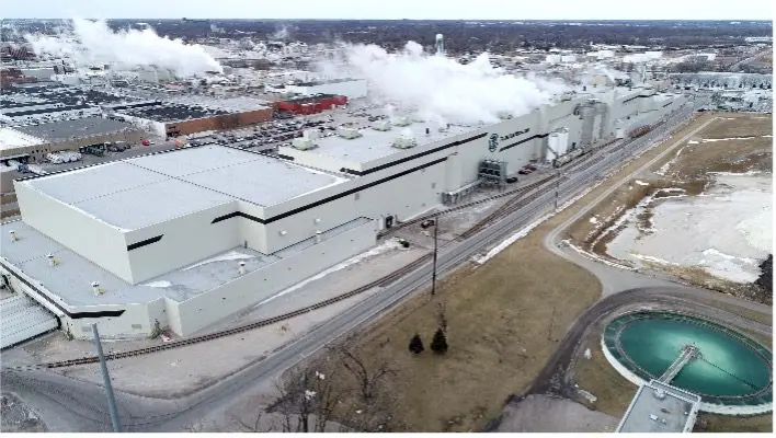 A large industrial facility with smoke rising from multiple sections, surrounded by roads, railway tracks, and a circular water basin, seen from an aerial perspective in a partially snow-covered landscape.