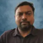 A man with short dark hair and a beard is wearing a dark collared shirt, posing against a blue, softly blurred background.