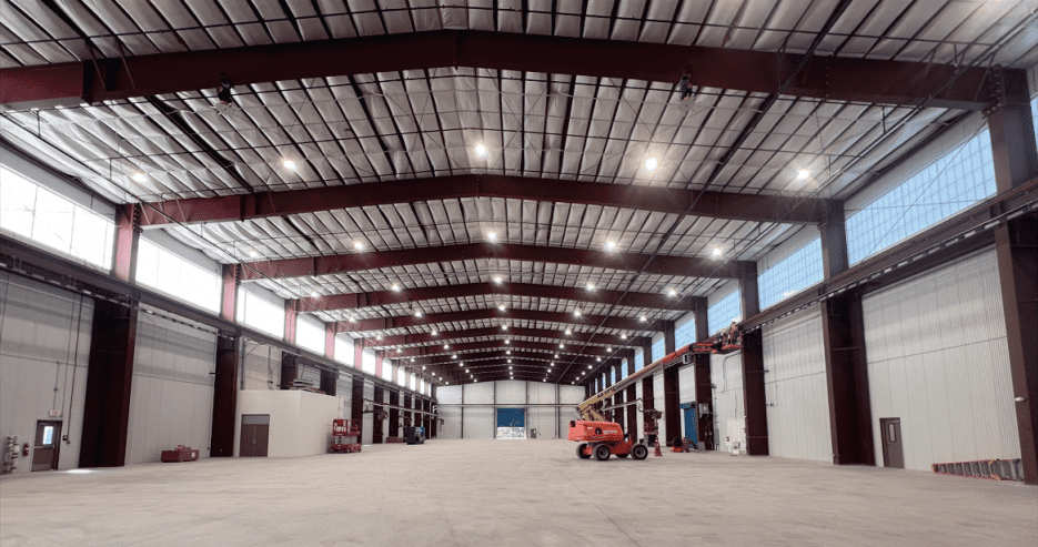 Spacious, empty warehouse with high ceilings, exposed steel beams, large overhead doors, and bright overhead lighting. A red boom lift is parked near one wall, and the concrete floor is clean and unobstructed.