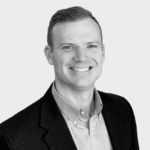 A man in a dark blazer and light button-up shirt smiles at the camera against a plain, light background.