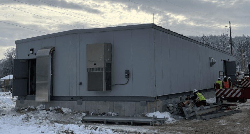 image001 (16) A large gray industrial building sits on a snowy construction site, with workers in safety vests and helmets working near stacked metal pipes and tools outside. Power lines and trees are visible in the background.