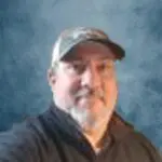 A man with a beard wearing a dark shirt and a baseball cap poses in front of a blue textured background.