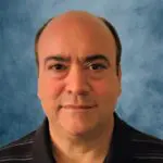 A middle-aged man with a receding hairline and short dark hair is wearing a black polo shirt with thin white stripes, standing in front of a blue textured background.