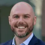 A smiling bald man with a beard, wearing a navy blue suit jacket and light blue shirt, stands in front of a blurred outdoor background.