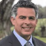A man with short, curly gray hair, wearing a dark suit, light blue shirt, and patterned tie, smiles while standing outdoors with greenery and blurred trees in the background.