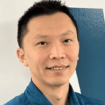 A man with short black hair, wearing a blue collared shirt, smiles at the camera in front of a white wall with a blue diagonal stripe.