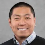 A person with short black hair, wearing small hoop earrings, a plaid collared shirt, and a dark sweater, smiles in front of a plain gray background.