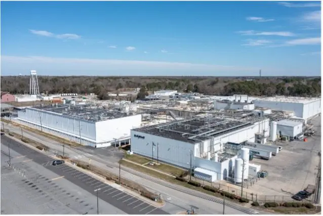 A large industrial facility with multiple white buildings and flat roofs, surrounded by empty parking lots and roads, with trees and a water tower in the background under a blue sky.