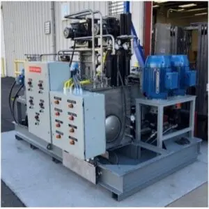 Picture1 Industrial hydraulic power unit with control panels, blue motors, and various pipes and wires, installed outdoors on a concrete pad near a metal building and an open warehouse door.