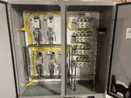 An open industrial control panel with wiring, switches, and metal pipes organized inside a gray metal cabinet. Yellow cables and electrical components are neatly arranged on both sides.