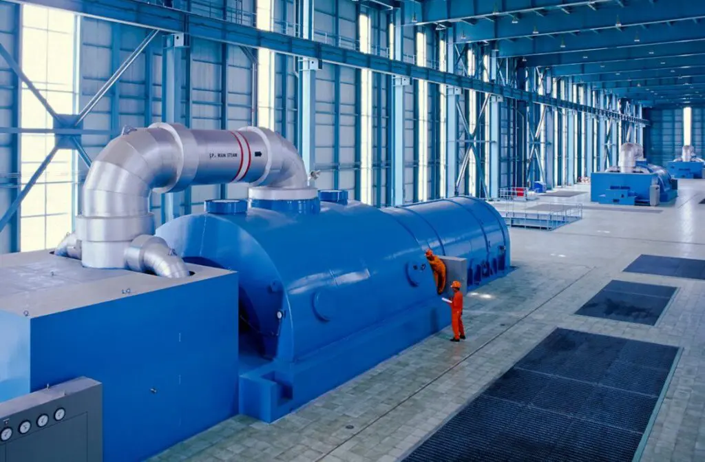image001 Two workers in orange uniforms inspect a large blue industrial turbine inside a spacious, modern, light-filled facility with steel beams and high ceilings.