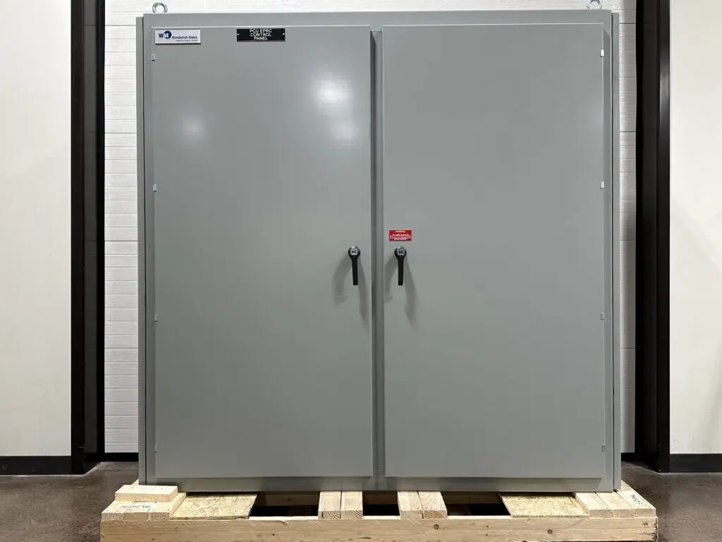 Photo 5-5-26, 1 43 19 PM A large gray metal cabinet with two doors and black handles stands on a wooden pallet indoors. Small labels and a warning sticker are visible on the cabinet’s upper corners and between the handles.