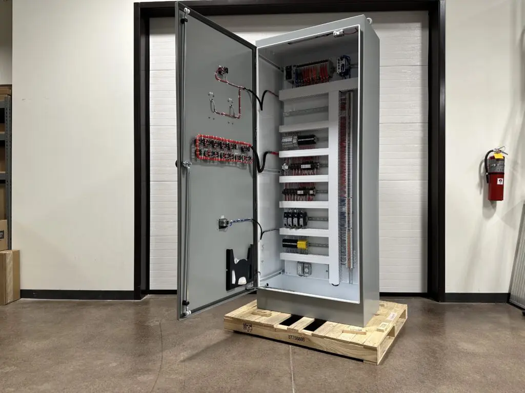 Photo 5-5-26, 1 54 59 PM A large electrical control panel with an open door showing internal wiring and components, standing on a wooden pallet indoors near a wall and a fire extinguisher.