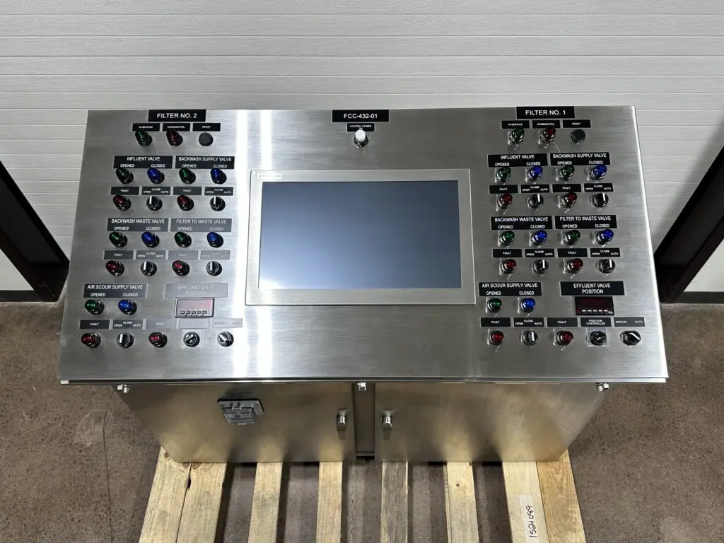 Photo 5-7-26, 1 21 28 PM A stainless steel industrial control panel with a central touchscreen display, surrounded by labeled buttons, switches, and indicator lights for operating and monitoring filter systems, placed on a wooden pallet indoors.