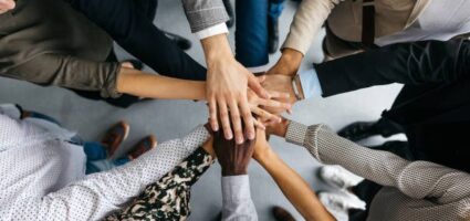 A high-angle shot of a group of male and female colleagues putting their hands together in an office. They are dressed in fashionable business clothes. Their faces are not visible, only their arms. Horizontal daylight indoor photo.