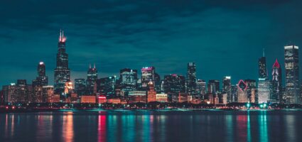 max-bender-8FdEwlxP3oU-unsplash Nighttime cityscape of Chicago with illuminated skyscrapers reflecting in the calm waters of Lake Michigan under a cloudy sky. The skyline features distinctive, brightly lit high-rise buildings.