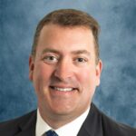 John David_Cutts A man in a suit and tie smiles in front of a blue textured backdrop. He has short brown hair and light skin.