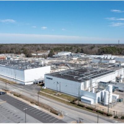 Picture1 A large industrial facility with multiple white buildings and flat roofs, surrounded by empty parking lots and roads, with trees and a water tower in the background under a blue sky.