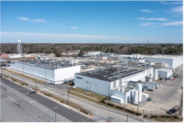 A large industrial facility with multiple white buildings and flat roofs, surrounded by empty parking lots and roads, with trees and a water tower in the background under a blue sky.