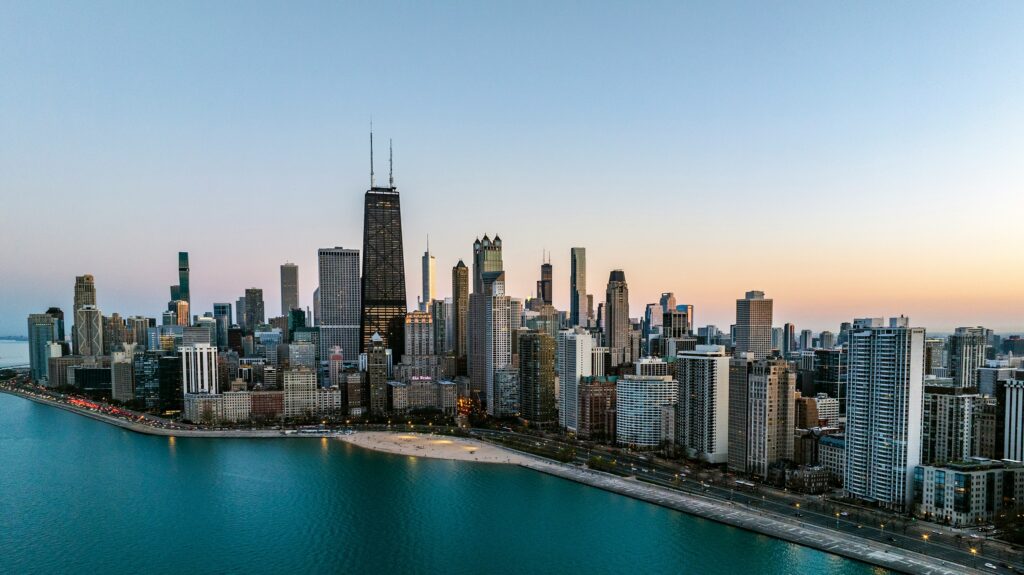 Aerial view of Chicago’s downtown skyline at sunset, with tall skyscrapers, including the John Hancock Center, along Lake Michigan’s waterfront. The sky is clear with soft evening light.