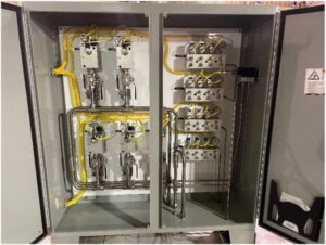A gray electrical control panel with an open door shows neatly arranged yellow wires, metal pipes, switches, and terminal blocks inside, displaying a well-organized industrial wiring setup.