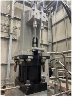 A close-up view of industrial machinery with metal rods, bolts, and a hydraulic or pneumatic actuator inside a factory setting with multiple pipes and cables in the background.