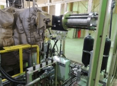 Industrial machinery with large hydraulic cylinders, metal pipes, and control valves inside a factory setting. The background shows yellow railings and padded equipment.