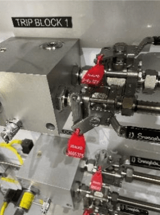 Close-up of an industrial valve system labeled "TRIP BLOCK 1," featuring metallic pipes and valves with several red security seals attached. Labels and yellow wires are visible in the background.