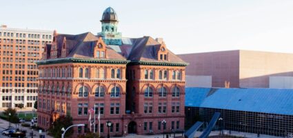 A historic red-brick building with arched windows and a green-domed tower stands next to a modern glass and metal structure on a city street with cars, trees, and flags visible.