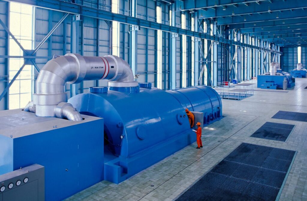 Two workers in orange uniforms inspect a large blue industrial turbine inside a spacious, modern, light-filled facility with steel beams and high ceilings.