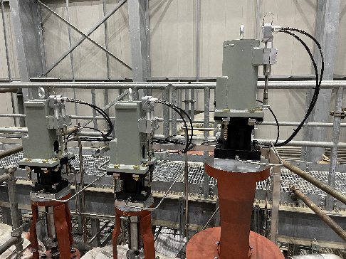 Three industrial control valves with attached actuators and cables are installed on red piping inside a facility with metal railings and a gray, open-framework background.