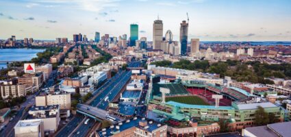 Aerial view of Boston, featuring Fenway Park in the foreground, busy roads, and the city skyline with tall buildings under a blue sky with scattered clouds.
