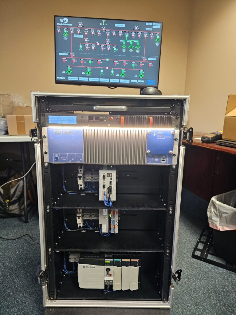 A rack-mounted industrial control system sits below a monitor displaying a network diagram with colored lines and icons. Various electronic modules and wiring are organized inside the open rack.