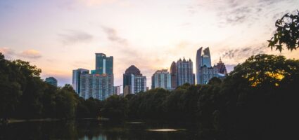 City skyline at sunset with tall buildings rising above dense trees, reflecting on a calm lake in the foreground. The sky is partly cloudy with soft pastel colors.