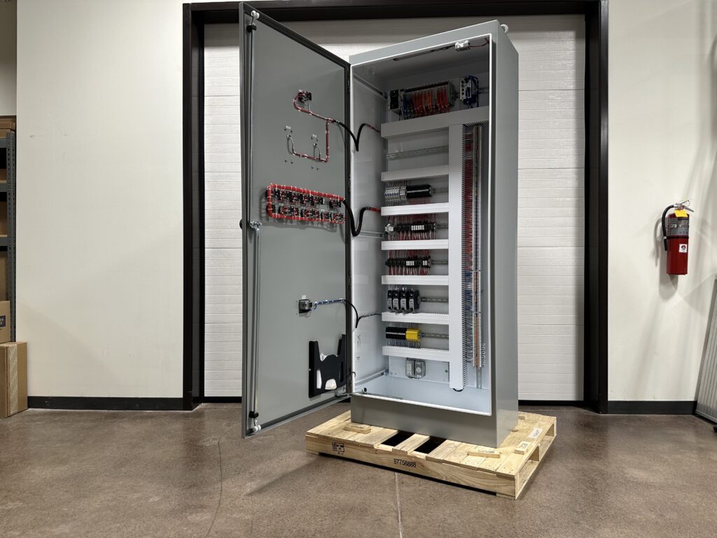 Photo 5-5-26, 1 54 59 PM A large electrical control panel with an open door showing internal wiring and components, standing on a wooden pallet indoors near a wall and a fire extinguisher.