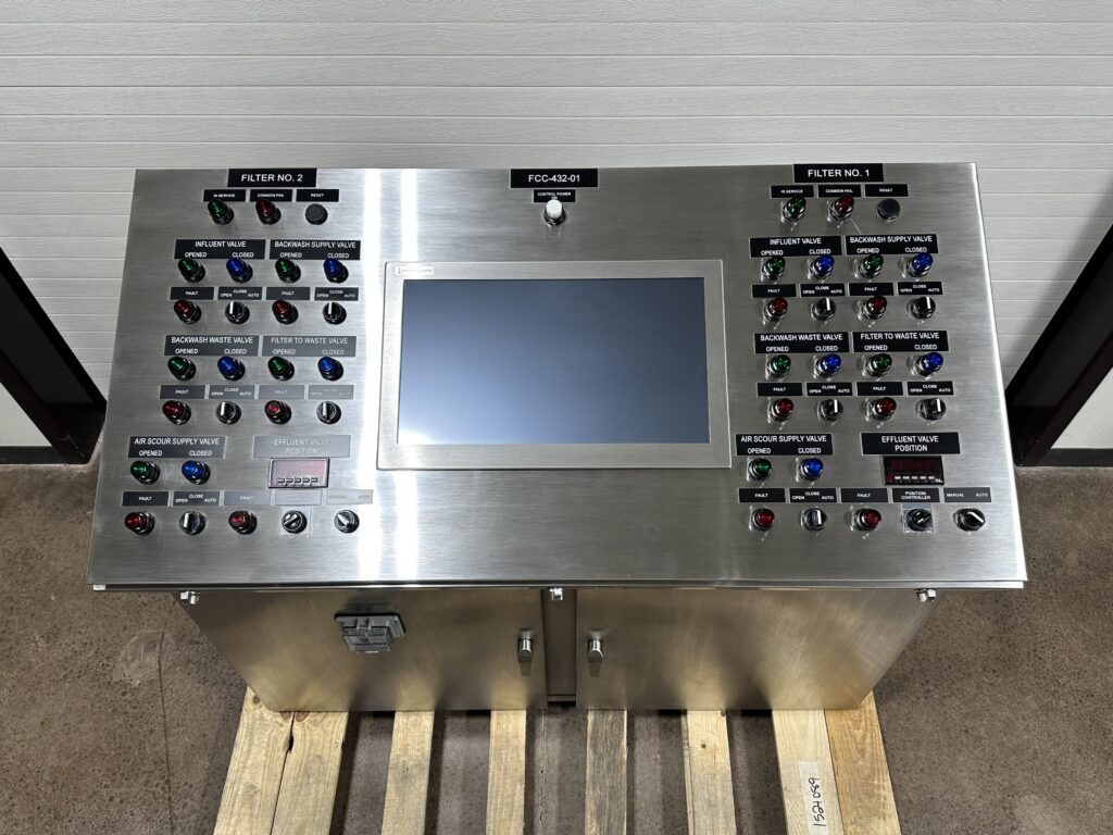 Photo 5-7-26, 1 21 28 PM A stainless steel industrial control panel with a central touchscreen display, surrounded by labeled buttons, switches, and indicator lights for operating and monitoring filter systems, placed on a wooden pallet indoors.