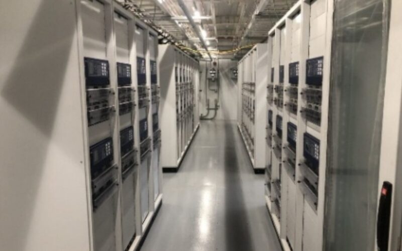 Screenshot 2026-05-26 at 1.58.39 PM A brightly lit server room with rows of white server racks on both sides, housing network equipment and cables, leading to the back of the room.